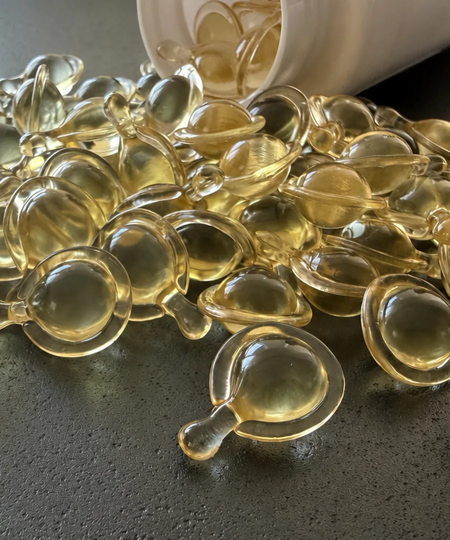 Plant-based single-dose twist-off capsules pouring from a white bottle — clear golden softgels for facial serum and skincare